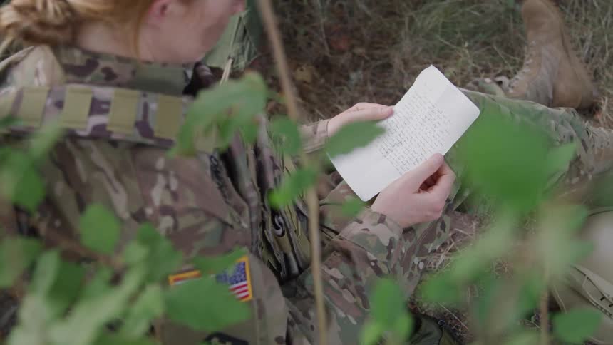 A woman in a camouflage uniform with a us flag patch reads a piece of paper while sitting outdoors amongst foliage