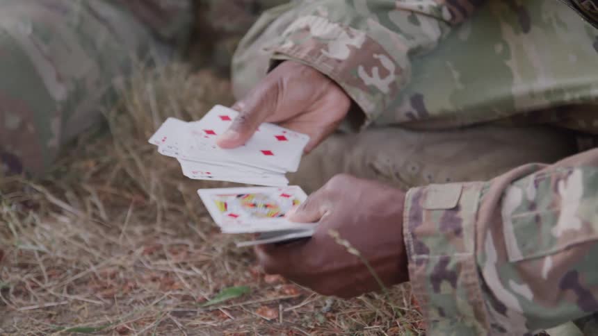 Hands wearing camouflage sleeves shuffle playing cards on dry ground outdoors