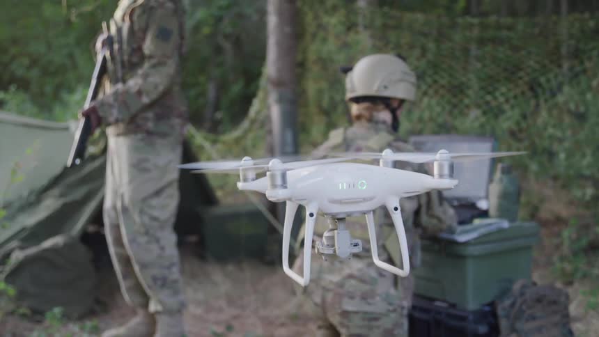 Two soldiers one holding a weapon and the other using a laptop with a drone appearing in the foreground