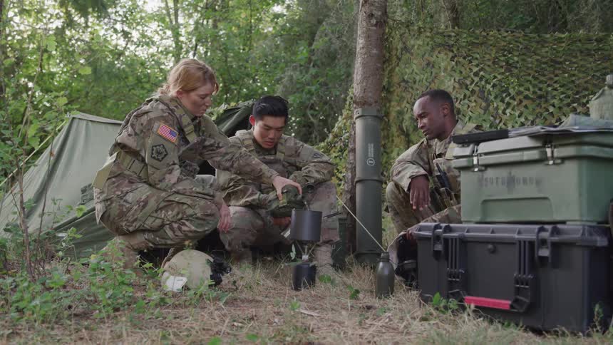 Two soldiers watch another female soldier pour water into a makeshift pot at a jungle camp