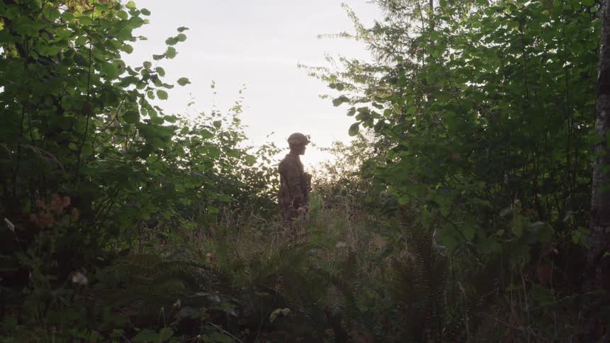 A soldier in camouflage clothing stands in a dense forest surrounded by various trees and plants
