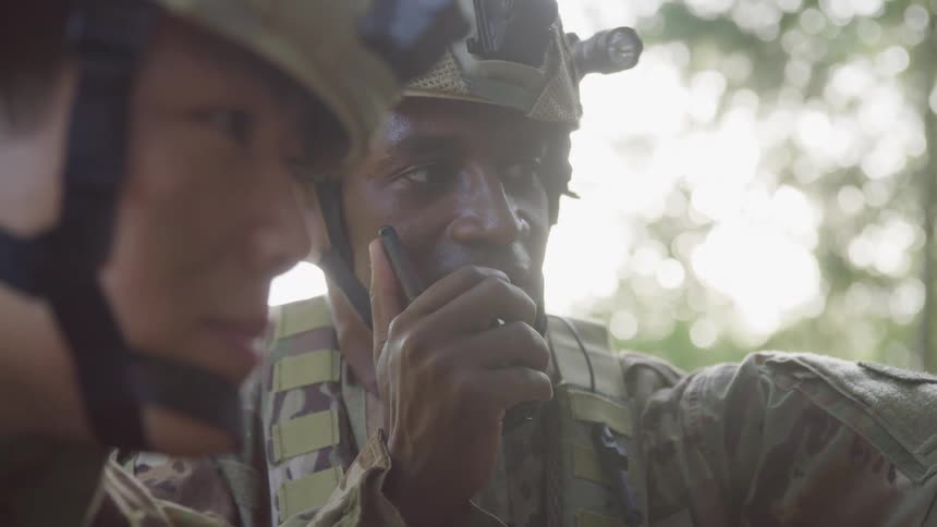 A person wearing a camouflage uniform and helmet speaks into a radio while looking at a screen in a wooded area with another individual nearby