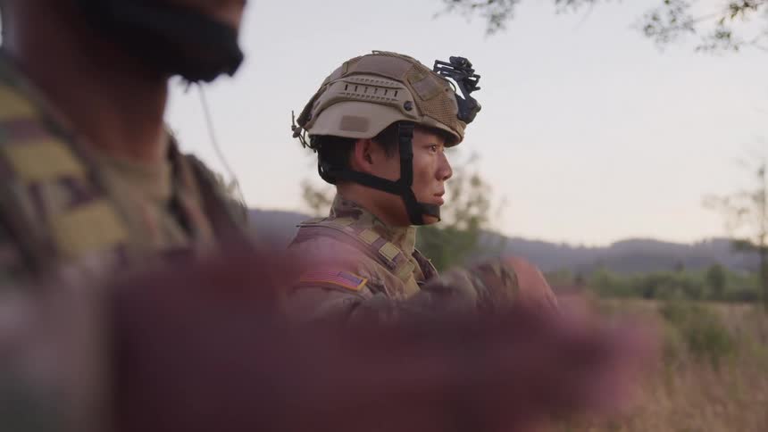 Two soldiers in camouflage uniforms and tactical gear are standing outdoors in a natural setting saluting