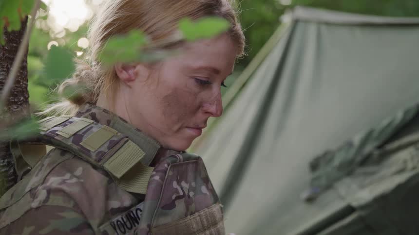Blonde woman in soldiers outfit leaning against a tree outdoors