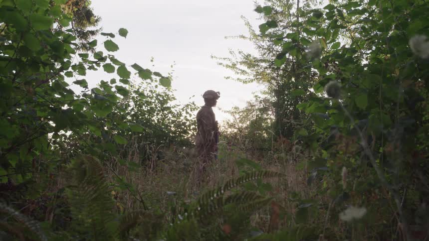 A soldier in camouflage clothing stands in a dense forest surrounded by various trees and plants