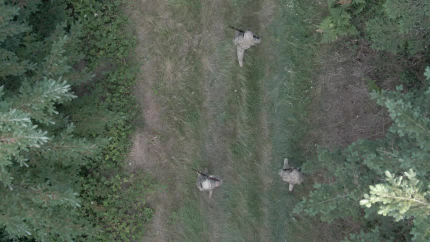 From a high aerial perspective three figures in camouflage clothing walking on a dirt path that cuts through a dense forest