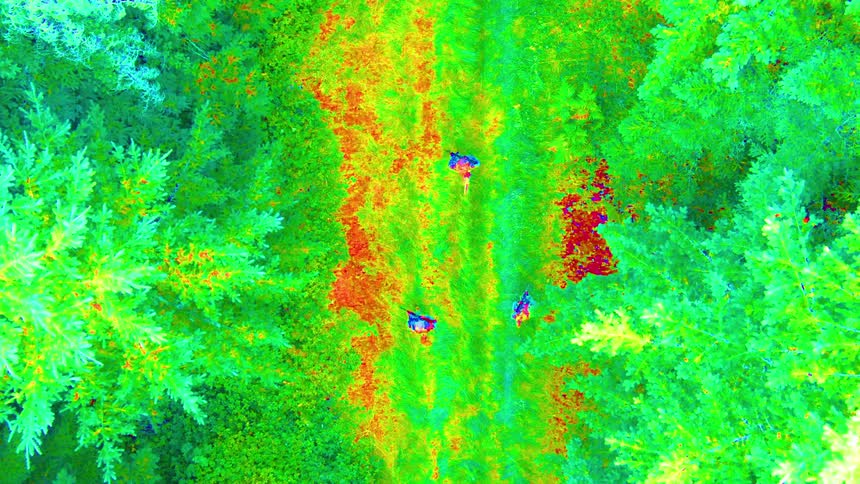 A falsecolor aerial thermal image displays a forested path with distinct heat signatures of military personnel moving through the terrain