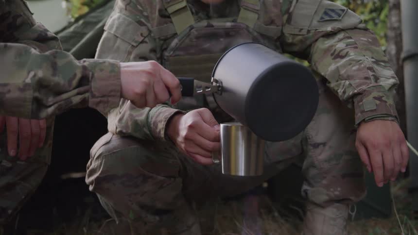 Two individuals in military uniforms are preparing food or drink outdoors with one person holding a metal cup and the other a pot
