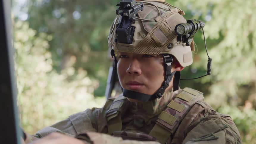 A soldier wearing tactical gear including a helmet with mounted night vision goggles and a communication device is seen in a forested area with another soldier in the background