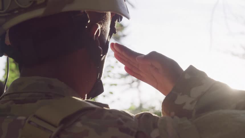 A soldier in camouflage uniform and helmet is saluting outdoors with sunlight streaming through trees in the background