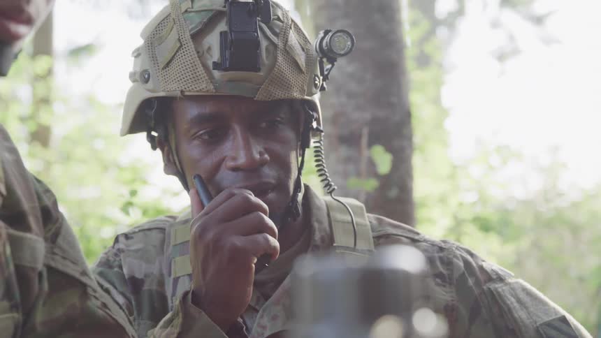 A person wearing a camouflage uniform and helmet speaks into a radio while looking at a screen in a forested area