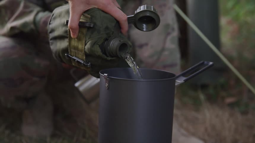 A person pours water from a militarystyle canteen into a dark metal pot outdoors while a camouflaged person crouches nearby