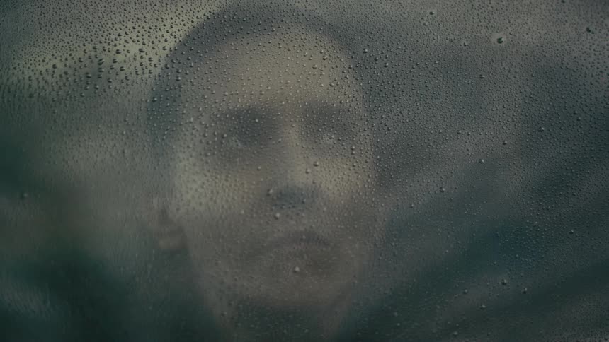 A persons face is seen through a foggy raincovered window