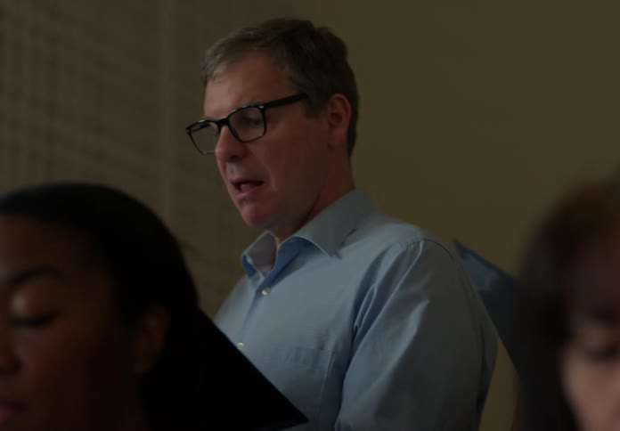 Two adult men one wearing glasses and a light blue shirt and the other a plaid shirt are shown looking downward singing in a choir in a softly lit room