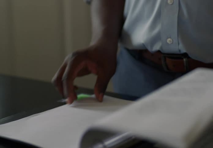 A black man wearing a gold wristwatch goes through a file in a folder flipping through the pages one by one