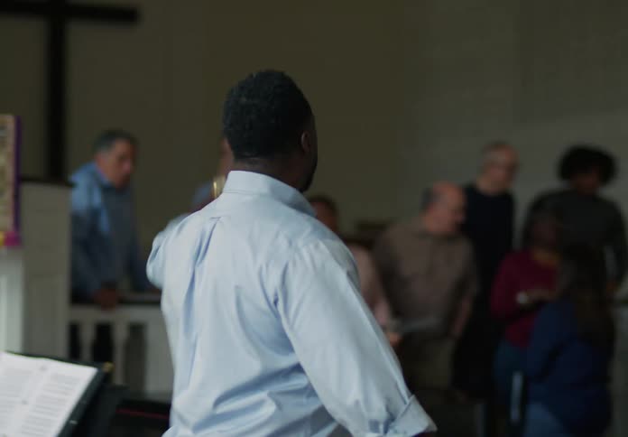 A man walks up to a group in a church who has hymnals in their hands ready to practice