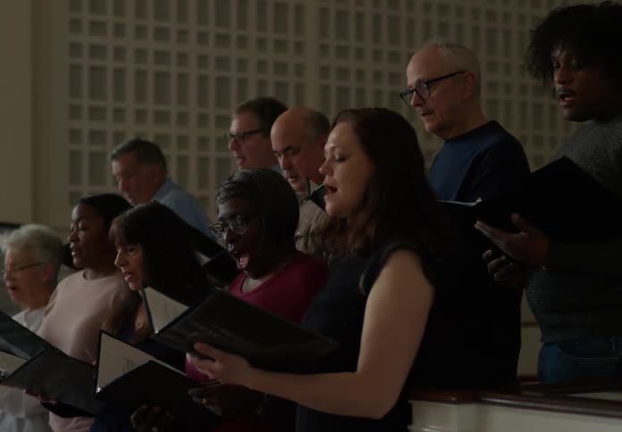 A group of people sings in a choir while a conductor sits nearby giving instructions