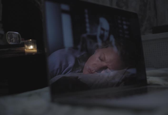 A person sleeps as a movie reflects on a laptop screen in a dim room