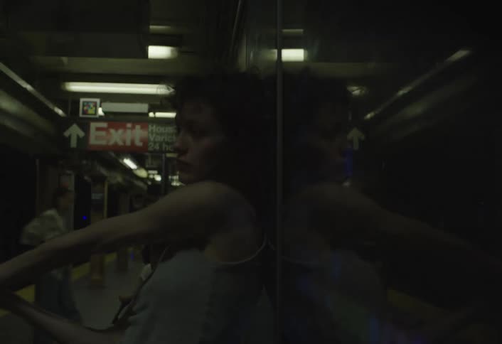A woman with curly hair leans against a reflective wall on a subway platform with an exit sign visible in the background