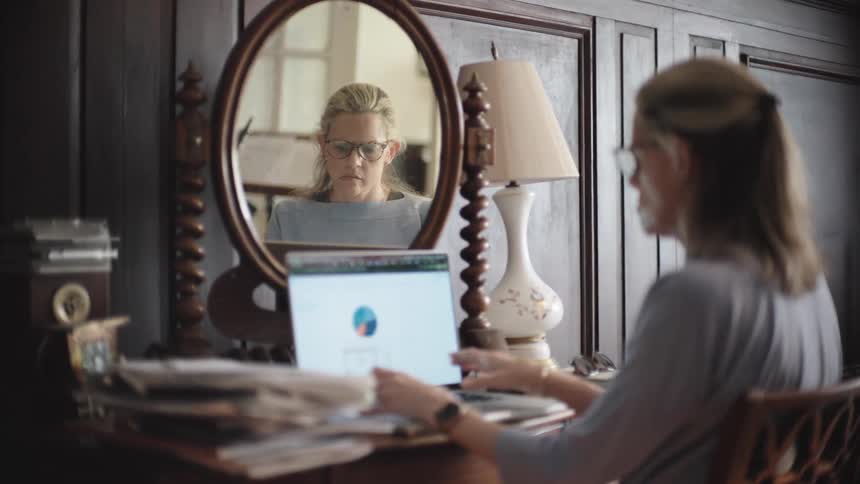 A woman works on a laptop at a desk with her reflection visible in a mirror behind the screen