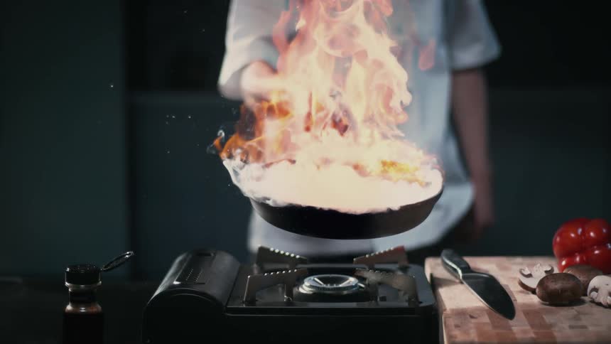 A chef holds a flaming pan over a burner