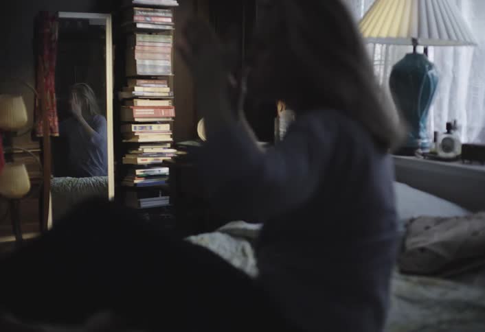 A woman in a dark shirt and pants wakes up in bed reaches for her head and sits up with a tall stack of books and a lamp visible in the background