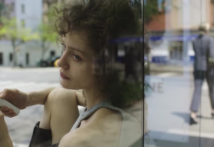 A woman with curly hair is seated beside a large window looking down with a city street visible in the reflection and background