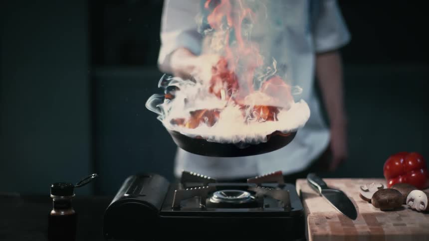 Person in kitchen holding flaming cast iron pan over stove