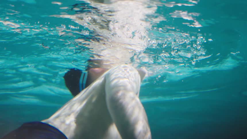 A child swims underwater in a swimming pool at midday wearing swimming goggles