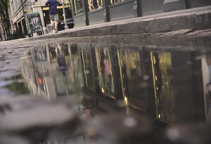 A person jogging in a city street is reflected in a large puddle on cobblestone pavement
