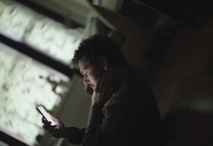 A man sits on the floor at an angle staring at a brightly lit phone screen in a dimly lit room