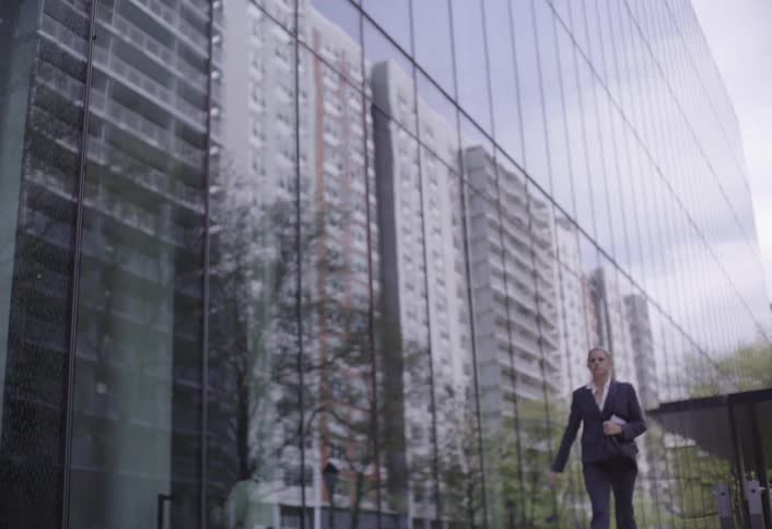 Woman in a pantsuit walks briskly past a building reflecting other nearby buildings during the daytime