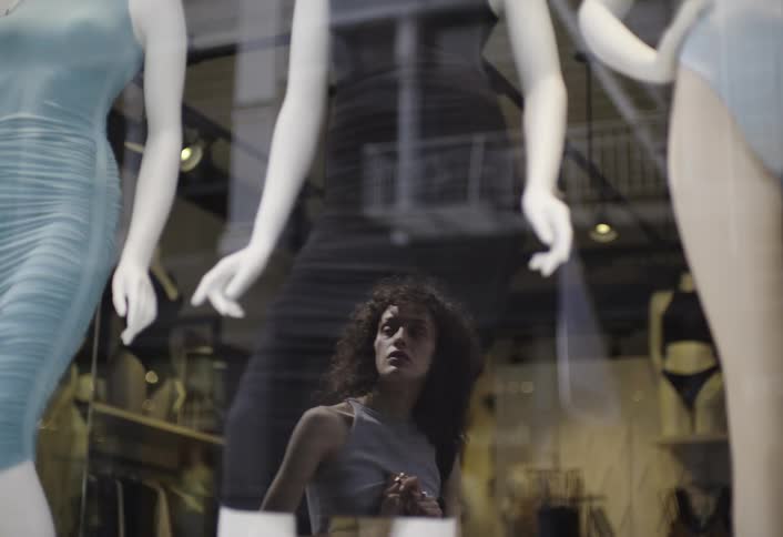A young lady stands outside a shop staring at the clothes worn by a mannequin through the glass