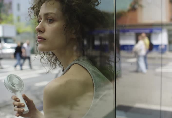 A woman sitting and leaning against a glass wall on the street holding a portable fan in her hand