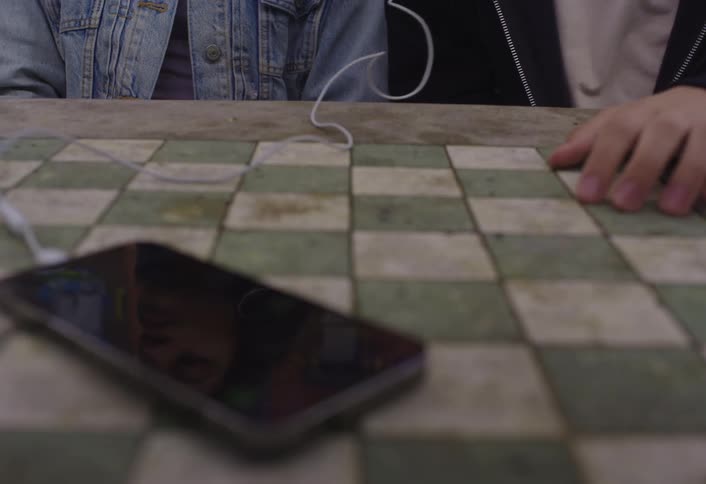 Two people sit at a checkered table sharing wired earphones connected to a phone