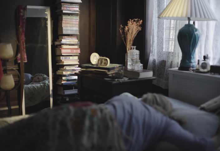 A woman lies on a bed facing away with her reflection in a fulllength mirror beside a tall stack of books then stands up