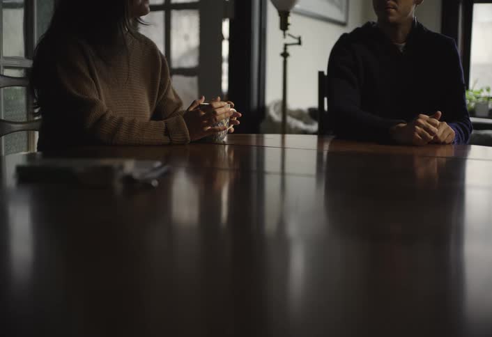 A man and a woman sit at a dining table engaged in a discussion with the woman holding a cup of coffee