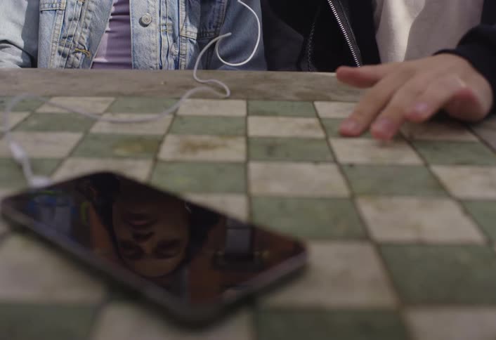 A smartphone on a table connected to headphones with two people sharing the headphones one of them is tapping their hand on the table to the rhythm of the music