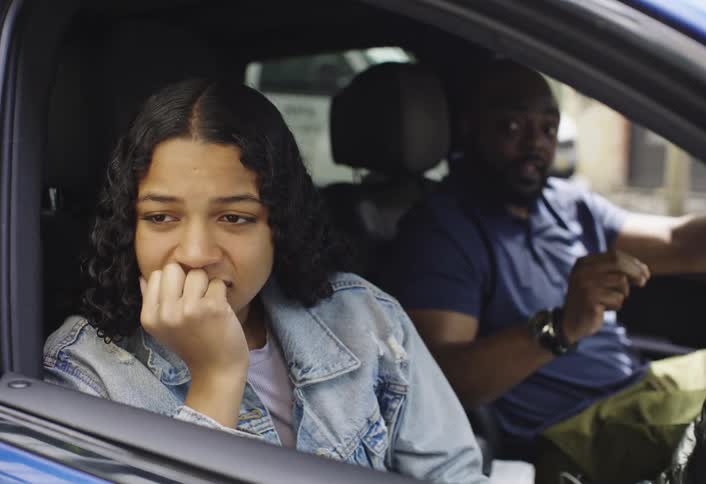 A teenage girl with dark curly hair sits in a car nervously biting their nails and looking down as an adult in the drivers seat reprimands her