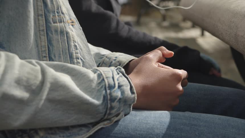 Two people sit side by side closely as one of them wearing a jean jacket rubs her fingers together