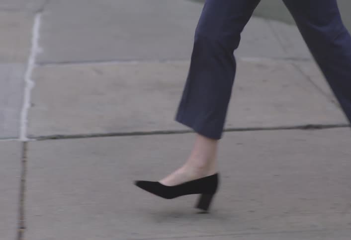 A person in blue trousers and black heels walks briskly across a sidewalk and then a crosswalk with a brief shot of their upper body adjusting their hair