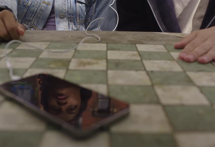 Two people sit side by side wearing wired earbuds connected to a smartphone on the desk listening to a song
