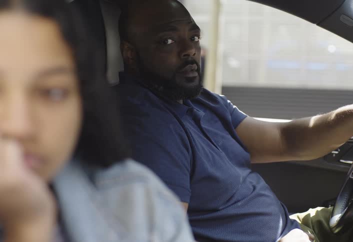 A man in the drivers seat glances seriously at a girl in the foreground inside a car