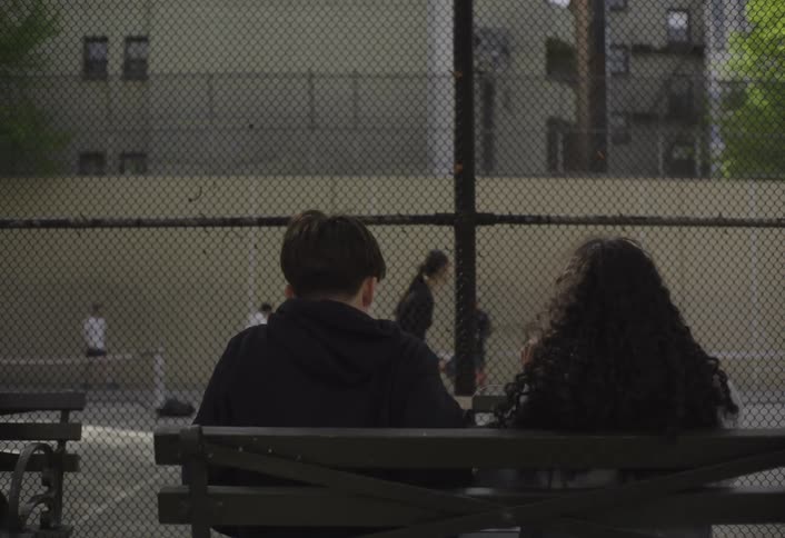 Teenagers share earbuds while sitting near a court others are playing pickle ball on