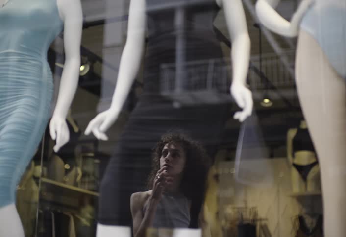 A girl stands outside on the street staring at a boutique store mannequin dressed in dresses