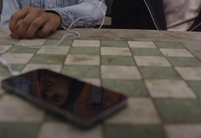 A phone sits on a table in front of two teenagers who are wearing and sharing a wired earbud
