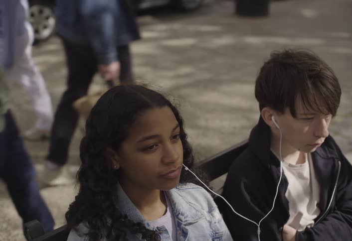 Young teenagers sharing earbuds sit on a bench on a windy day while pedestrians walk by in the background