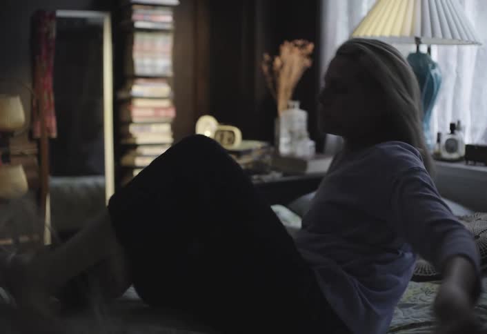 A person lies under a floral comforter on a bed wakes up startled and stands up with a stack of books her reflection in a mirror and a bedside lamp visible in the room