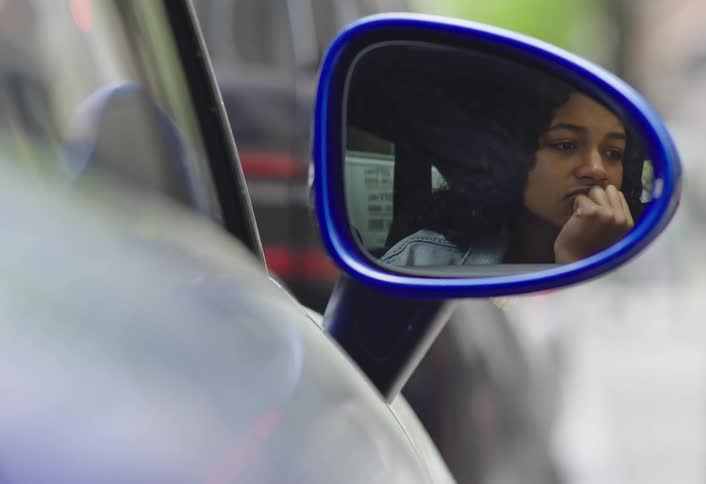 A thoughtful woman is reflected in a blue framed car side mirror resting her chin on her hand as she looks outside