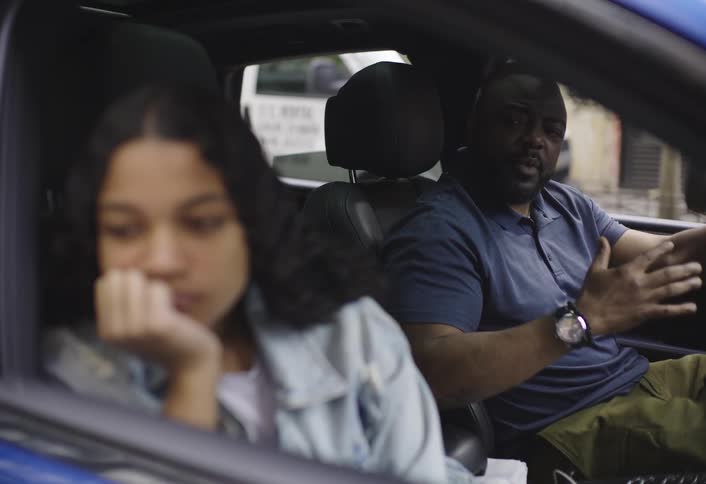 A blurred young woman sits pensively in the passenger seat of a car while the man in the drivers seat actively speaks and gestures appearing distressed